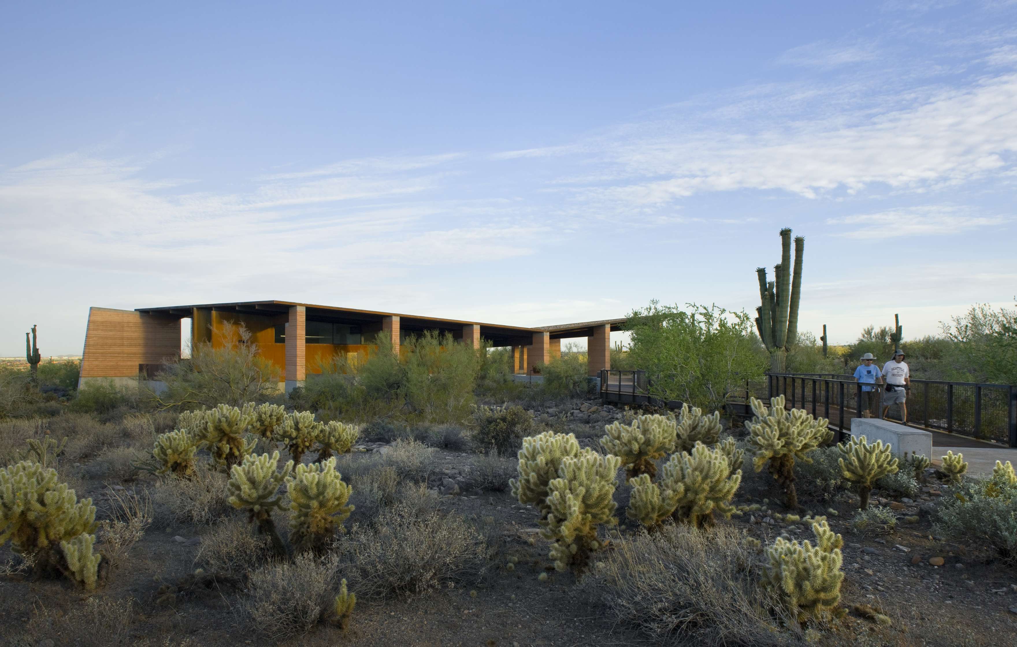 Three people walk on a path near a modern building surrounded by desert vegetation, including cacti, against a backdrop of a clear blue sky.