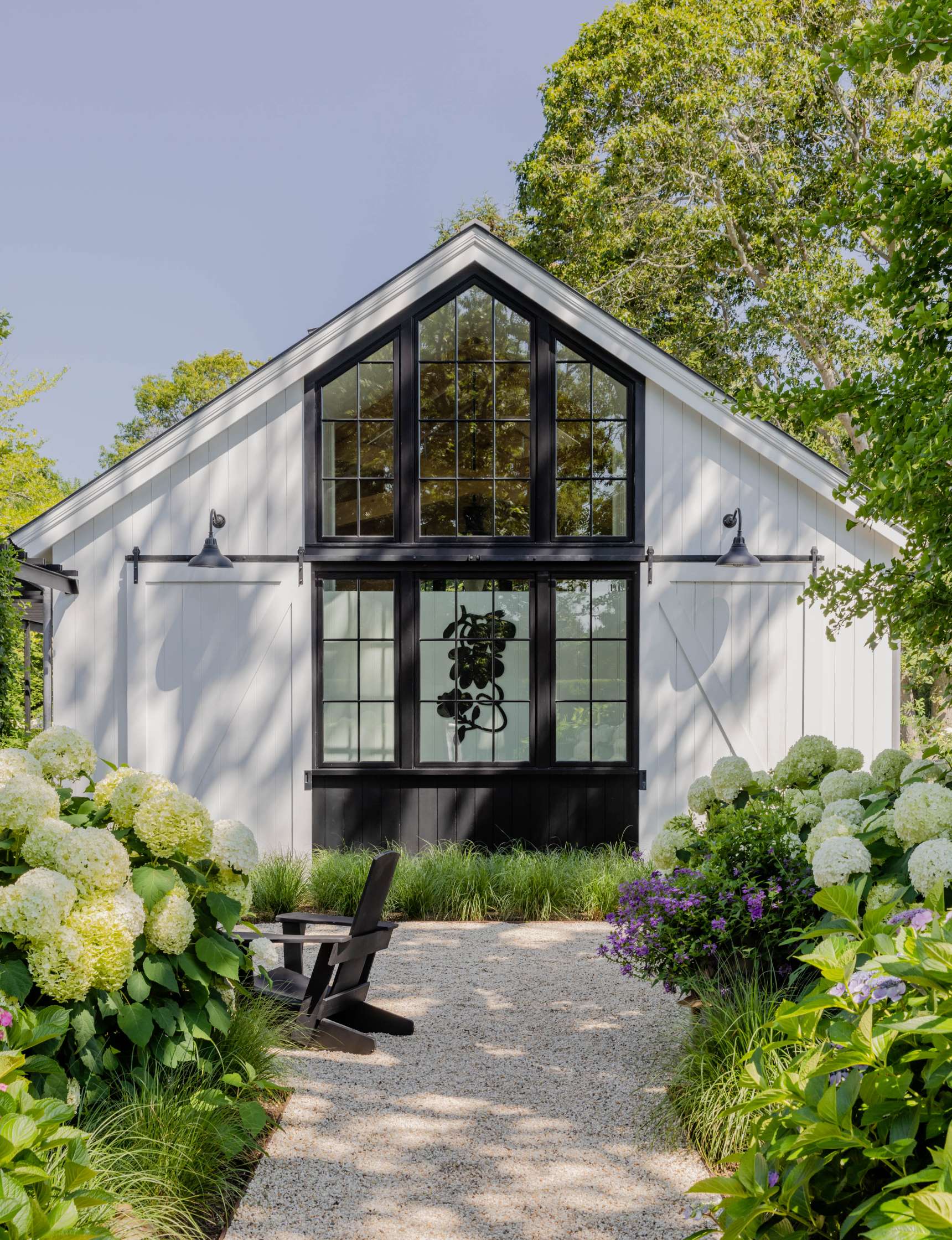 A white barn-style house with black window frames, surrounded by hydrangeas and greenery. Two black chairs on a gravel path lead to the entrance.