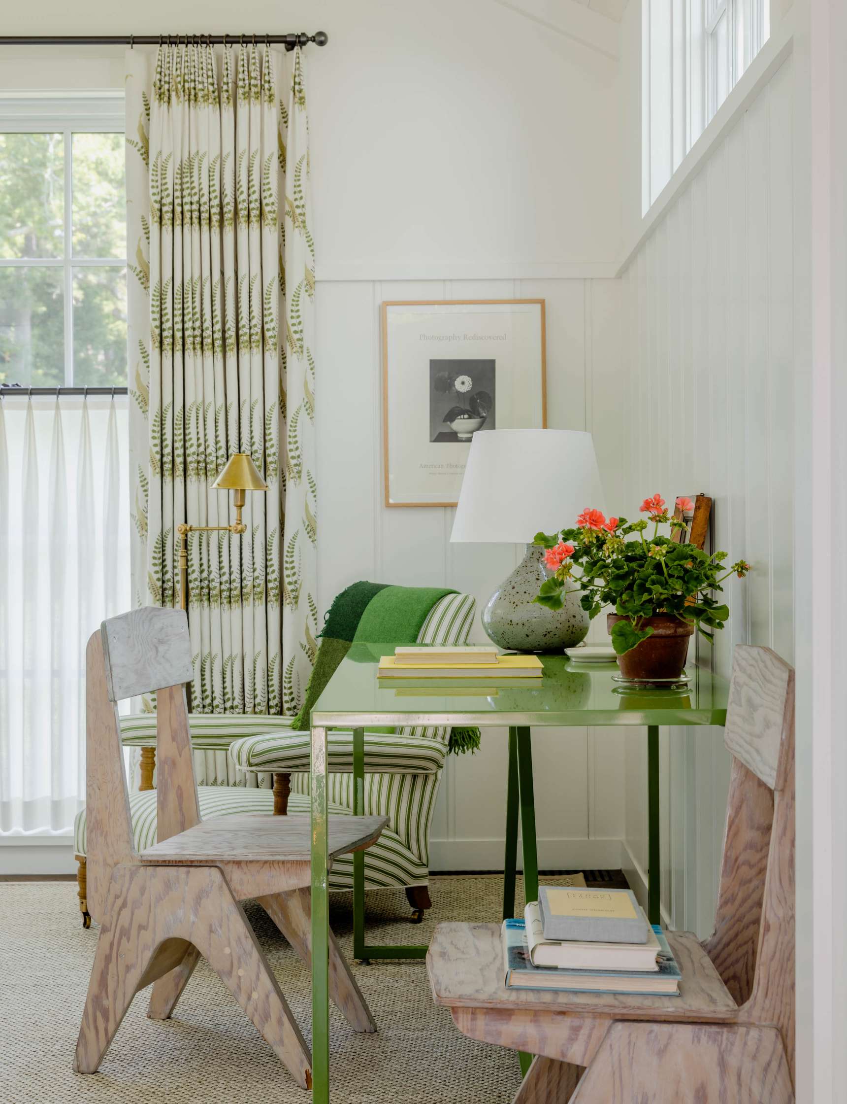 A small, brightly lit room features a green table with a lamp, books, and plants. Two wooden chairs sit by the table, and a striped armchair is partially visible. Large windows have white and patterned curtains.