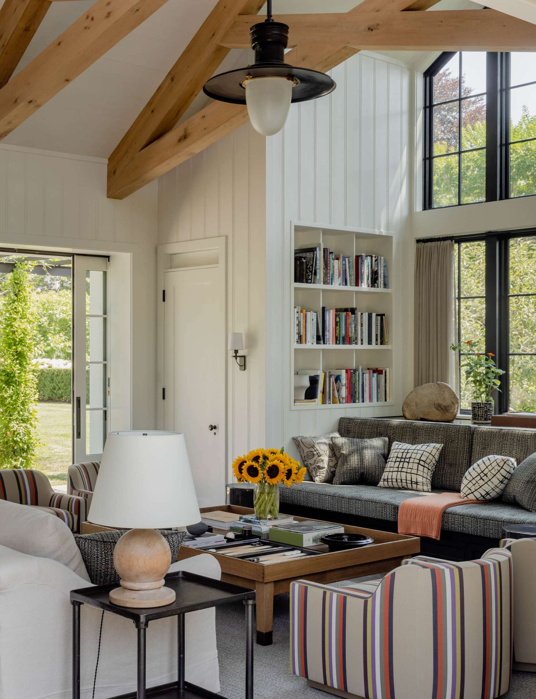 A cozy living room features a gray sofa with striped cushions, a coffee table with books and sunflowers, and a white armchair. Exposed wooden beams and a wall-mounted bookshelf add charm.