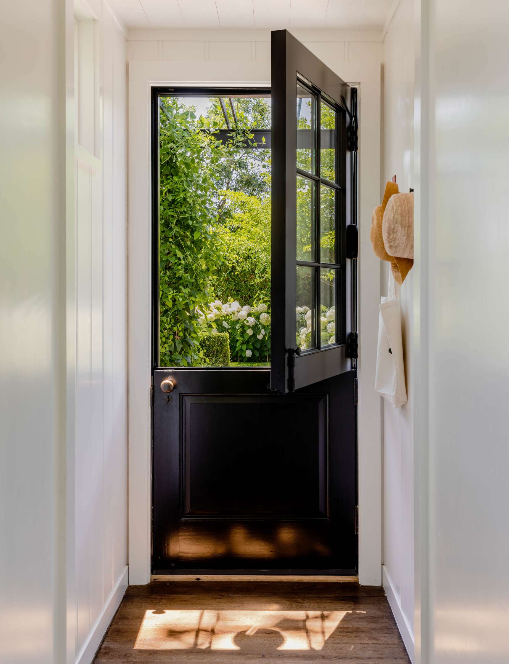 A half-open black Dutch door with a glass upper panel leads to a lush garden view. Two hats and a bag hang on hooks on the right wall of a narrow white hallway.