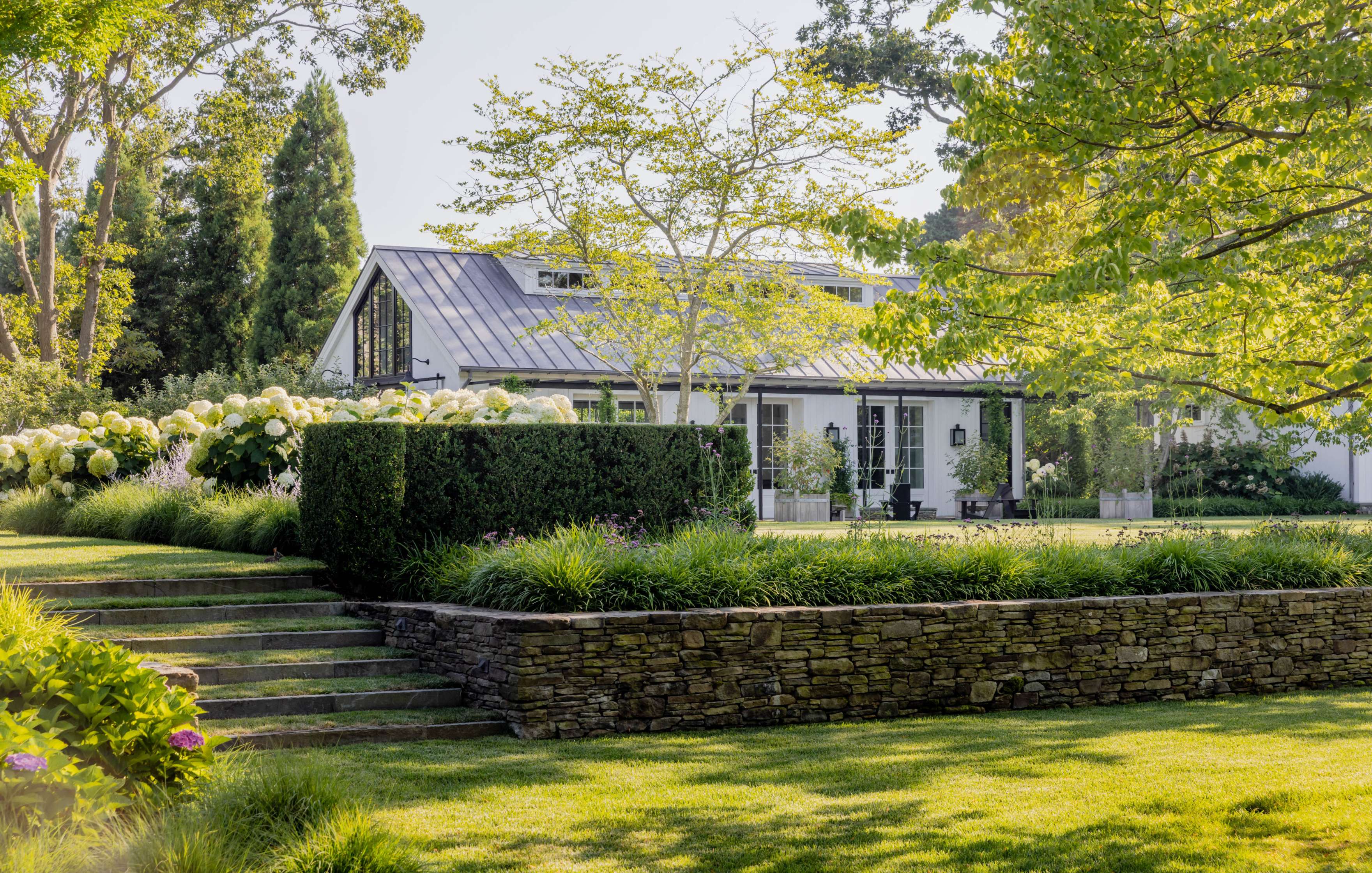 A white house with large windows and a metal roof is surrounded by trees, bushes, and a well-maintained lawn with stone steps leading up to it.