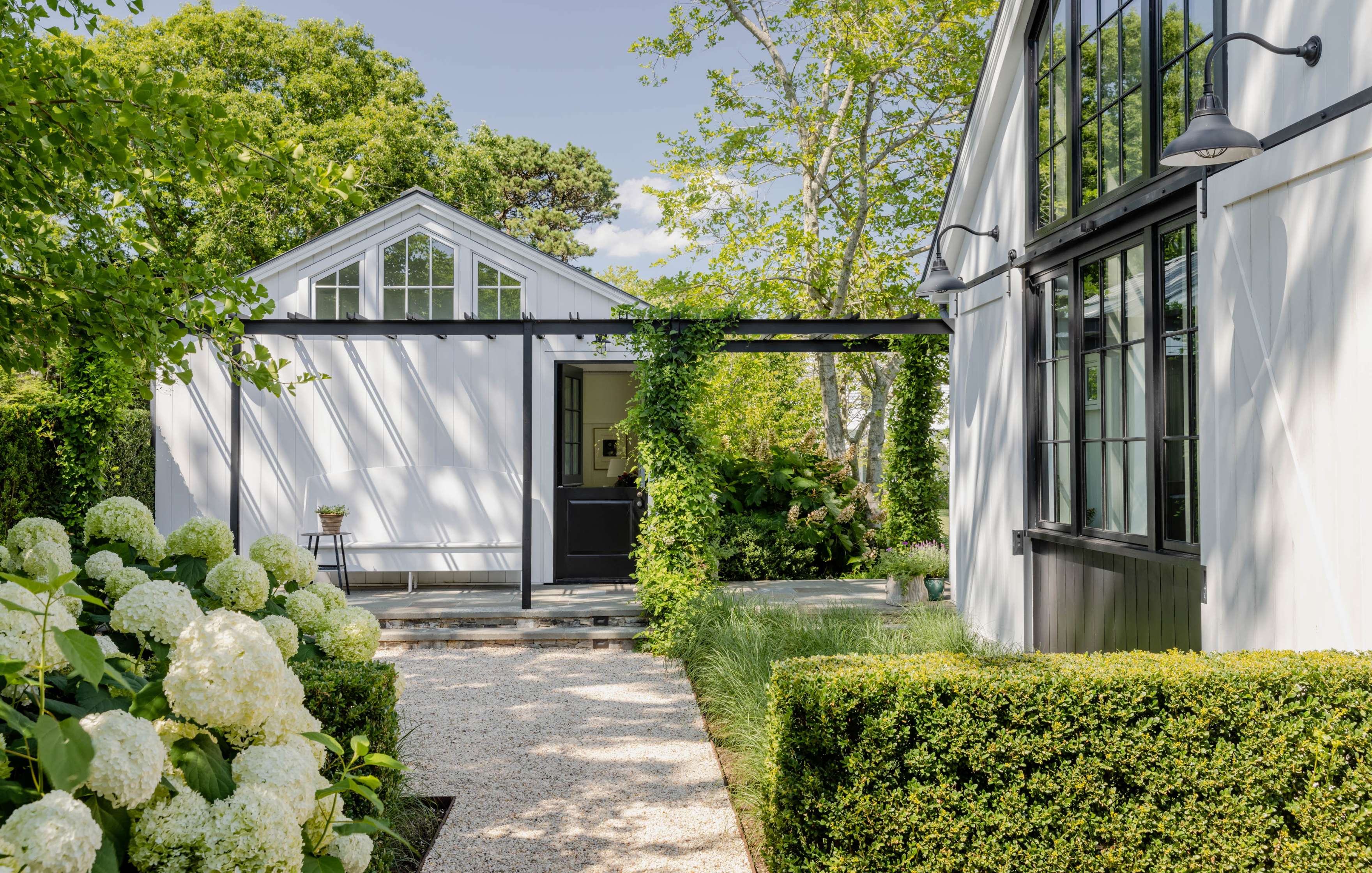 A modern white house with black window frames and a connecting pergola, surrounded by lush greenery and blooming white hydrangeas.