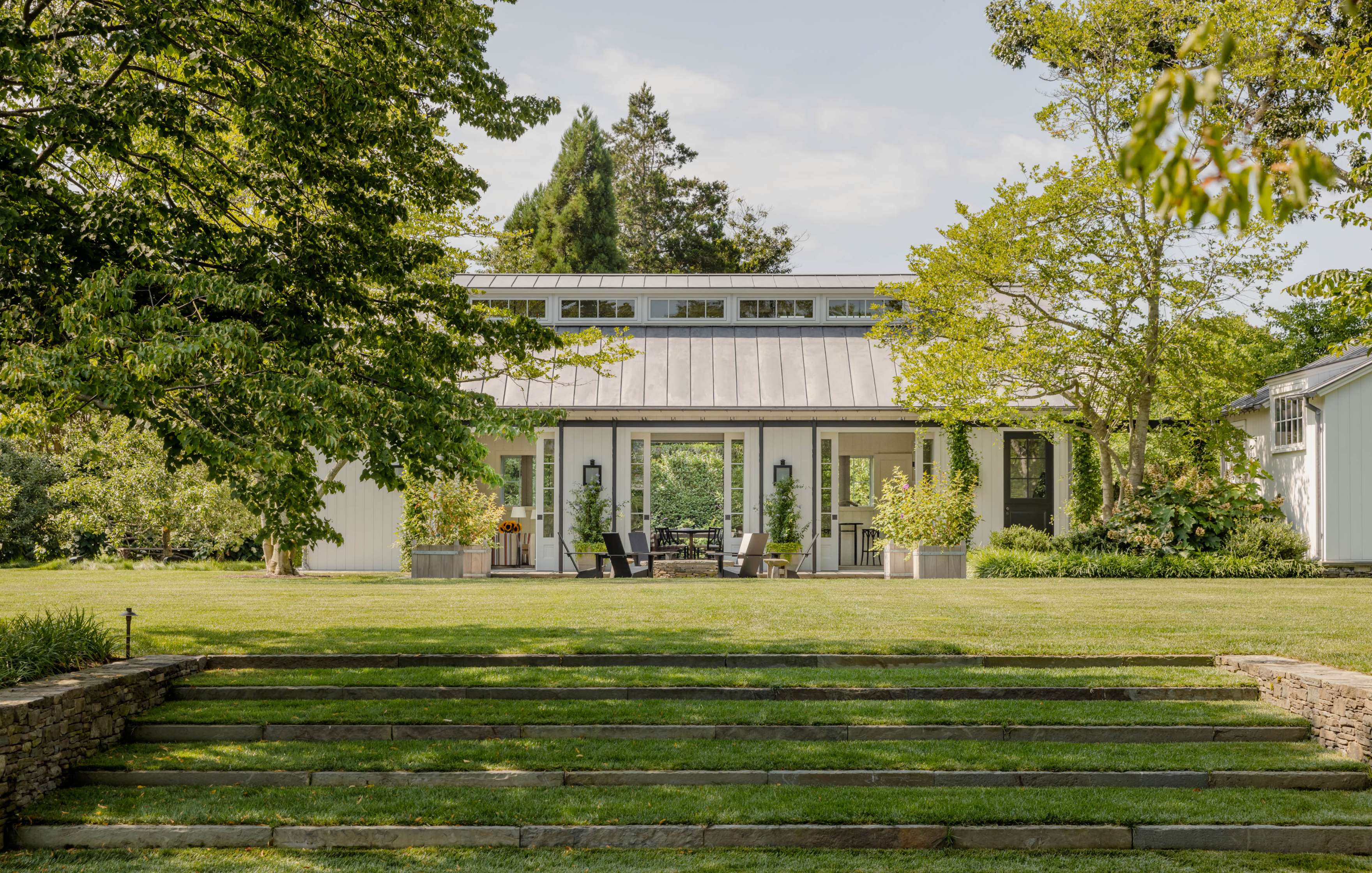 A modern white house with large windows and a metal roof is surrounded by trees. In front, there are two black chairs and potted plants on a well-manicured lawn with stone steps leading to the house.