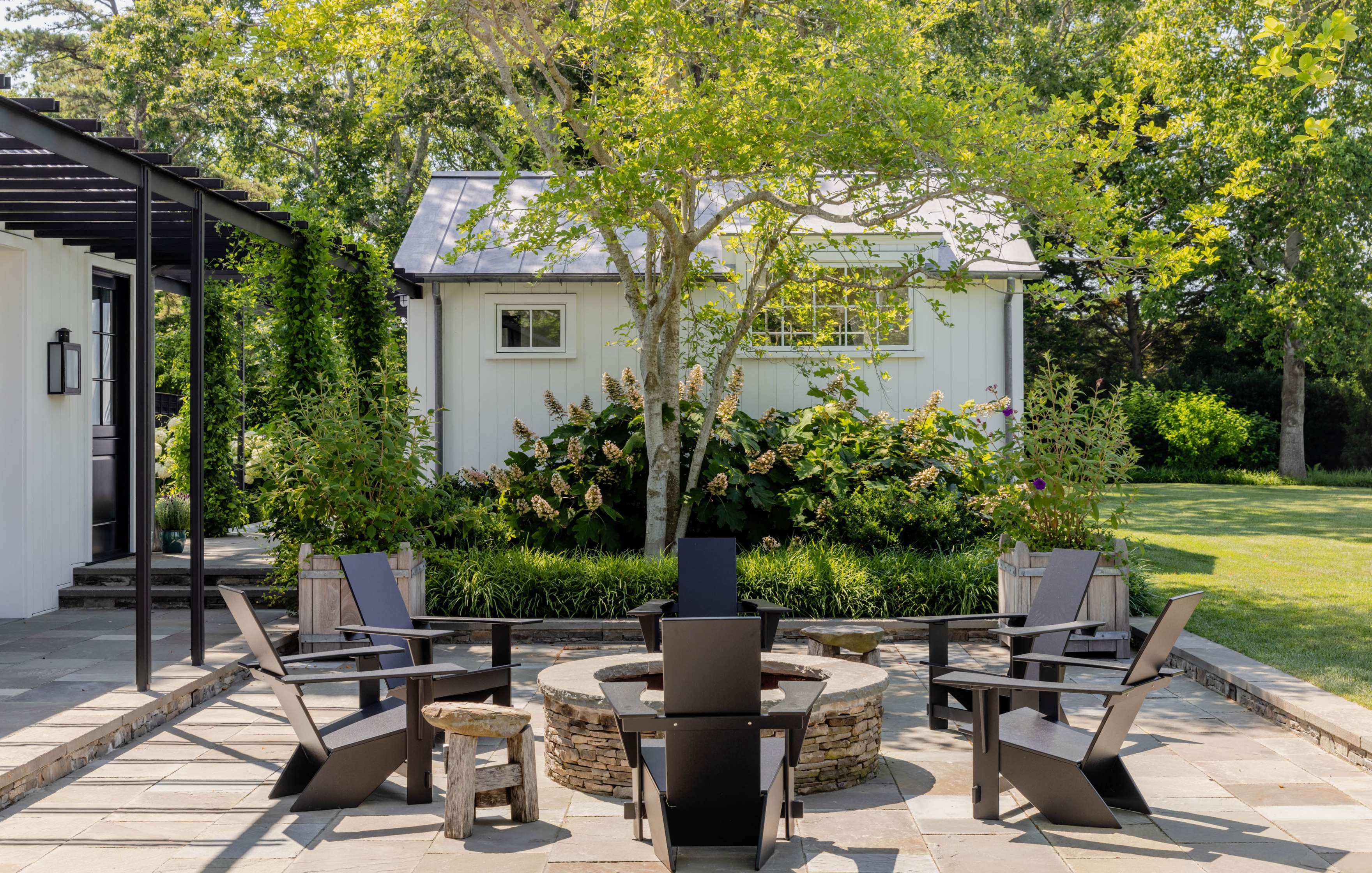 Outdoor patio with dark modern chairs arranged around a circular fire pit. A small white shed is in the background with greenery and a tree surrounding it.