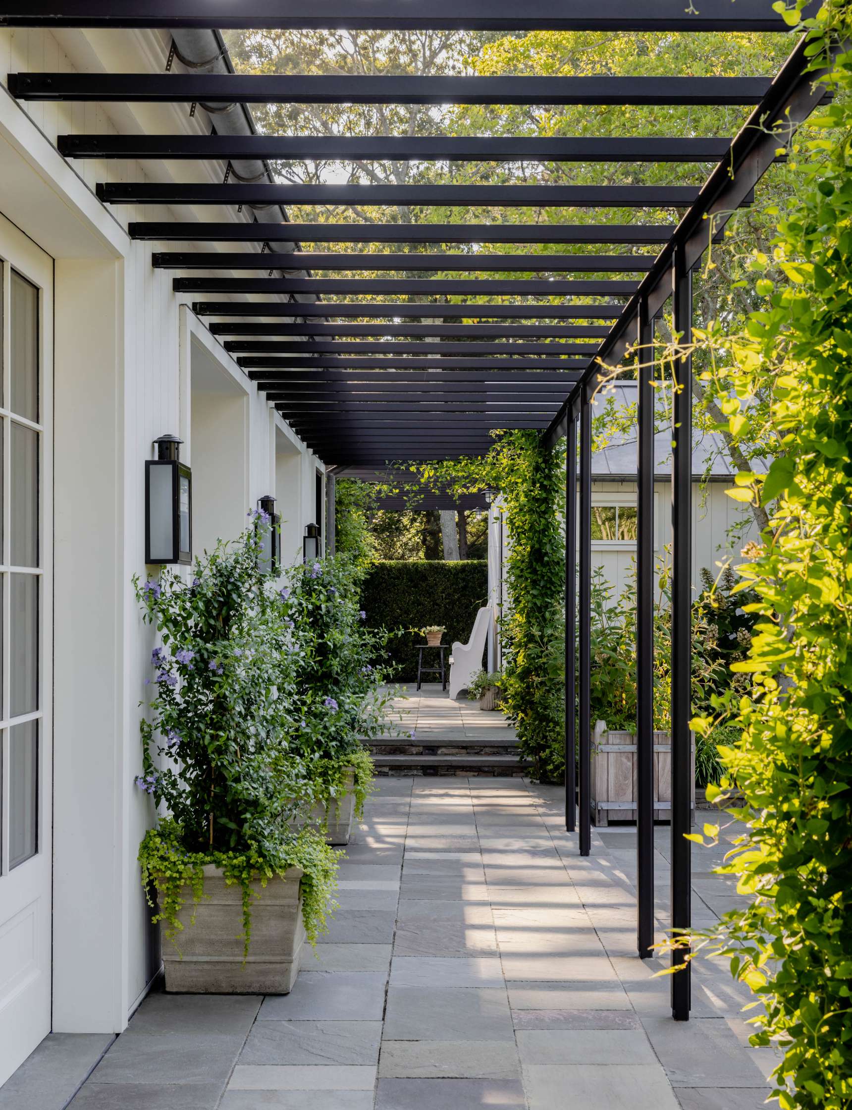 A paved walkway with potted plants on the sides is shaded by a black pergola structure attached to a white building. Lush greenery surrounds the area.