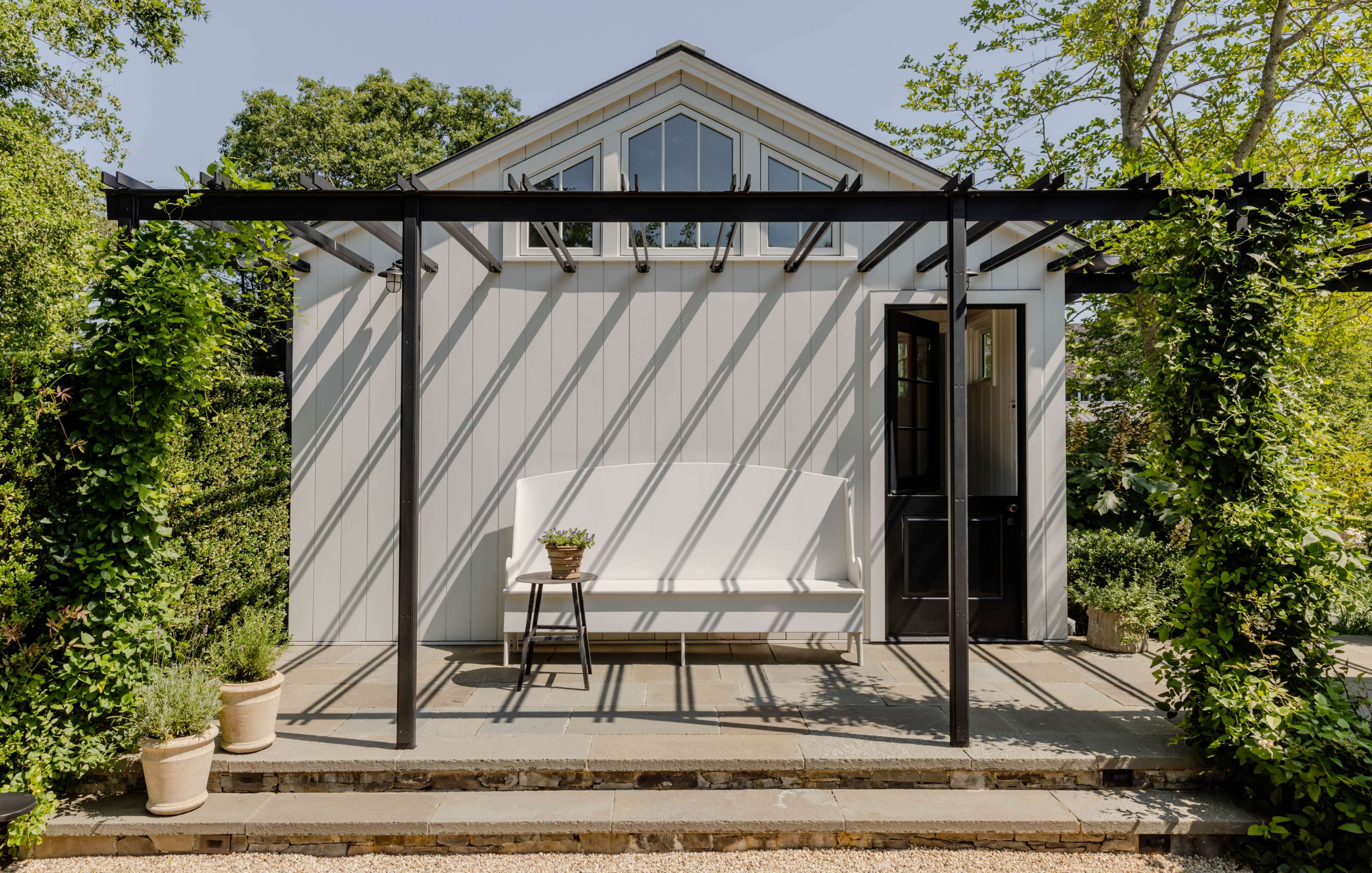 A small white building with a black-framed door and windows, featuring a covered patio area with a white bench and a plant on a side table. Two potted plants are placed in the foreground.