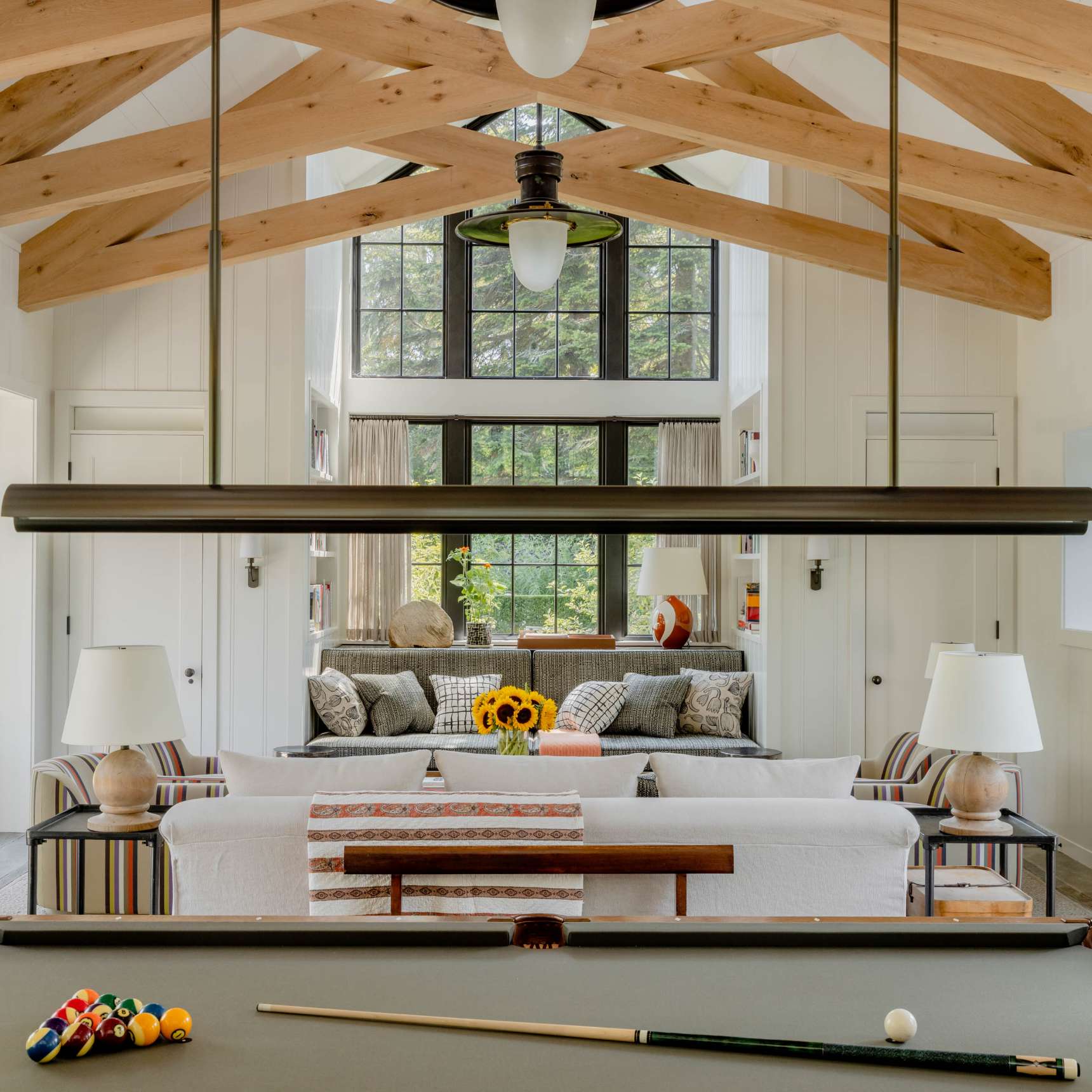 A spacious living room with a pool table in the foreground, a white sofa and two table lamps, large windows, and wooden ceiling beams. Sunflowers sit on the coffee table.