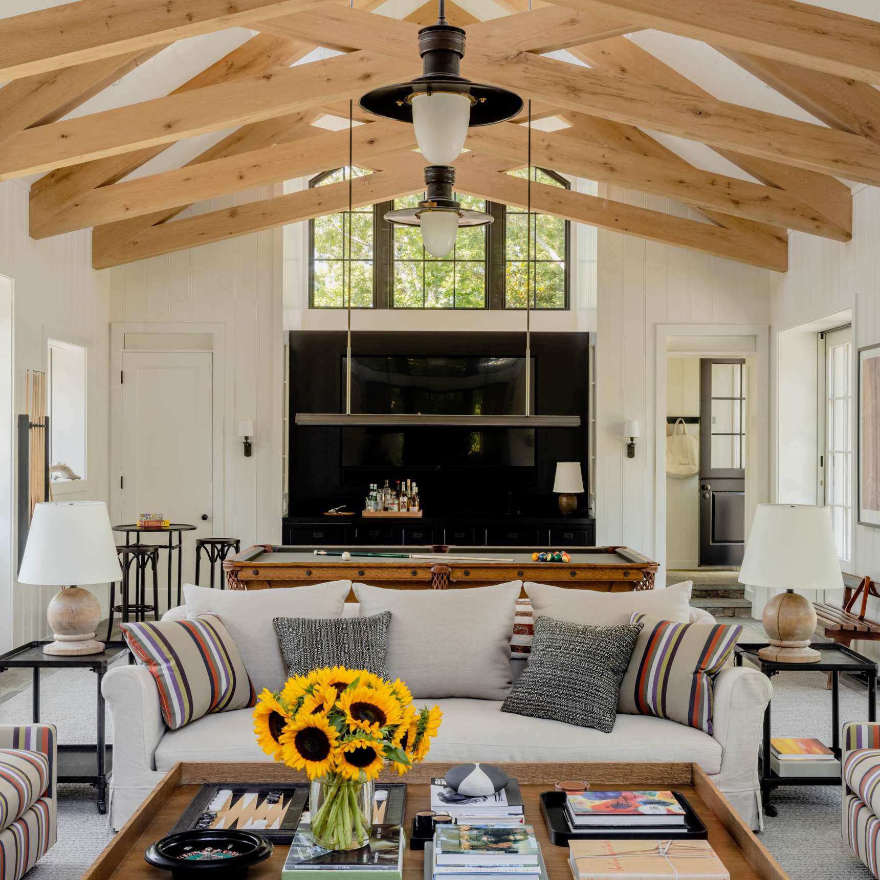 A well-lit living room with wooden beams, a beige sofa with striped arms, sunflowers on the coffee table, and a large TV screen on the wall. Natural light streams through tall windows.