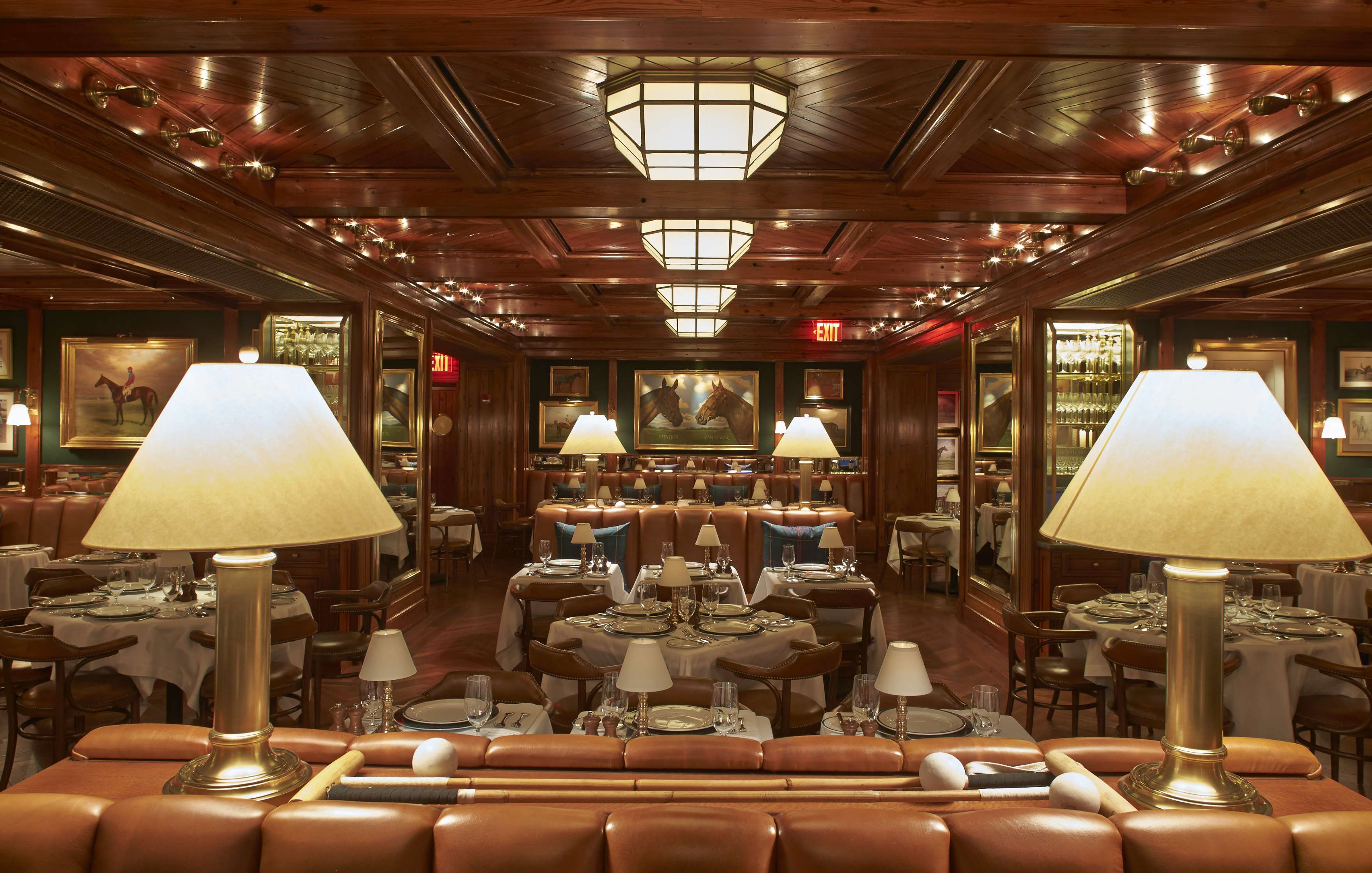 A dimly lit restaurant with wooden ceiling, beige leather seating, tables set with white tablecloths, and large lamps on the ledge in the foreground. Paintings adorn the walls.