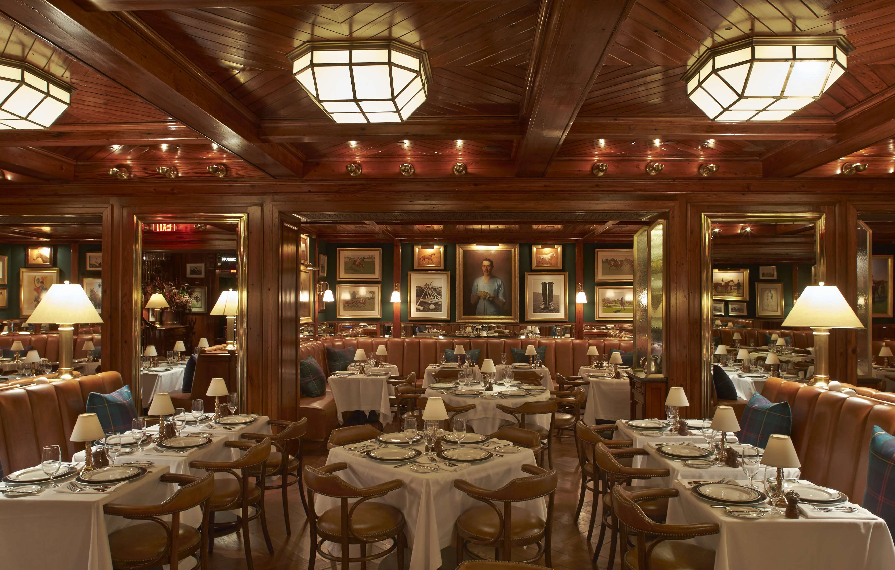 A dimly lit restaurant with wooden interiors, set tables with white tablecloths, brown chairs, and framed artworks on the walls.