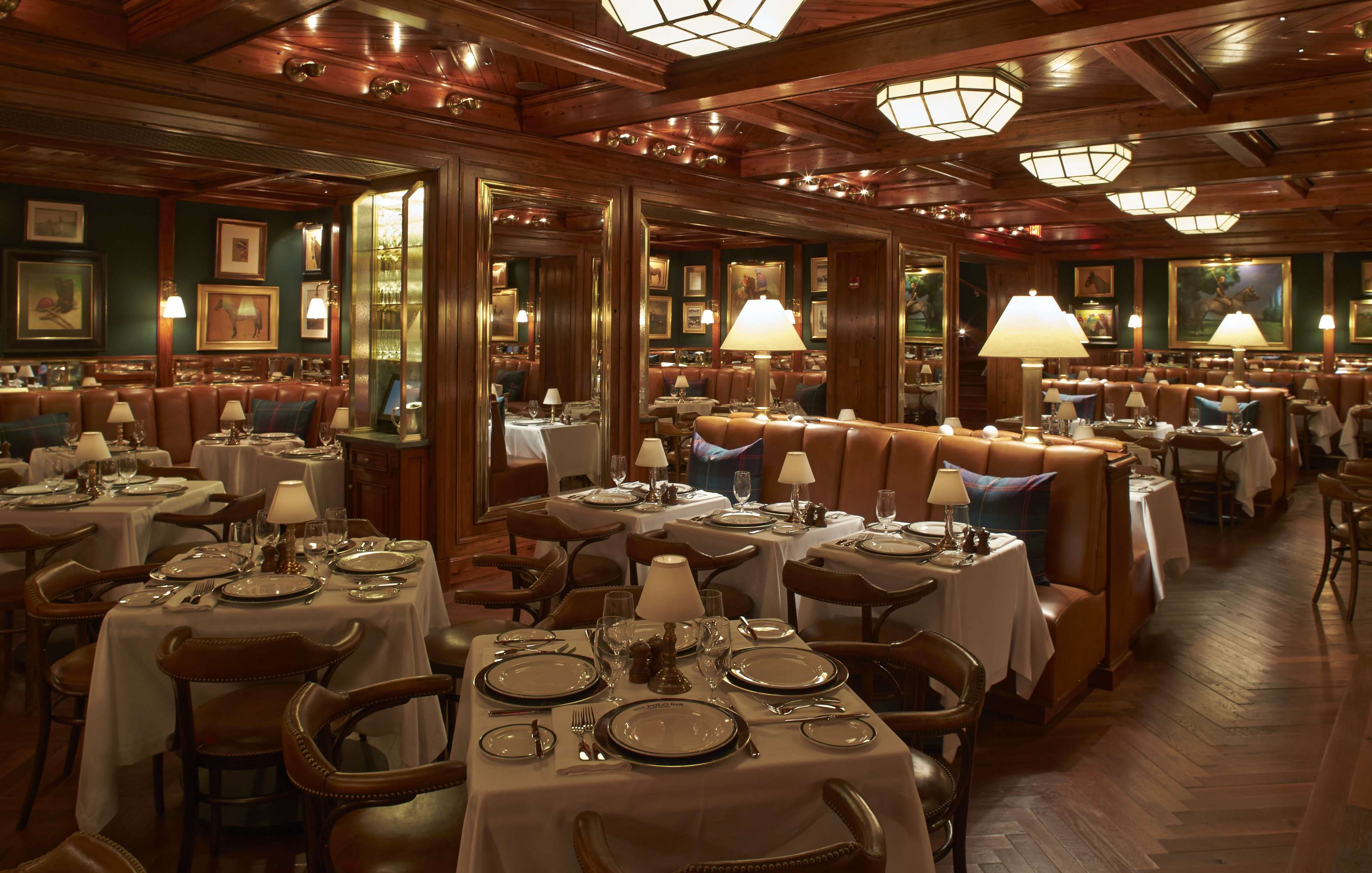 Elegant restaurant interior with wooden decor, leather seating, white tablecloths, and lamps on each table. The room is well-lit and has framed pictures on the walls.