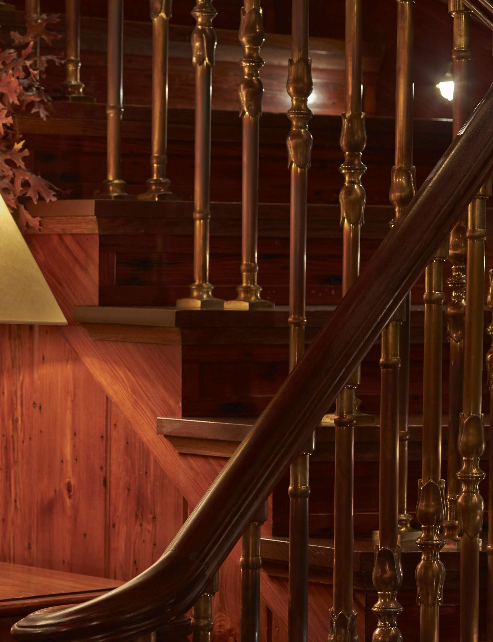 Wooden staircase with brass balusters, accompanied by a wooden handrail and a lit lamp on the left side. In the background, a wall is partially decorated with leaves and wood paneling.